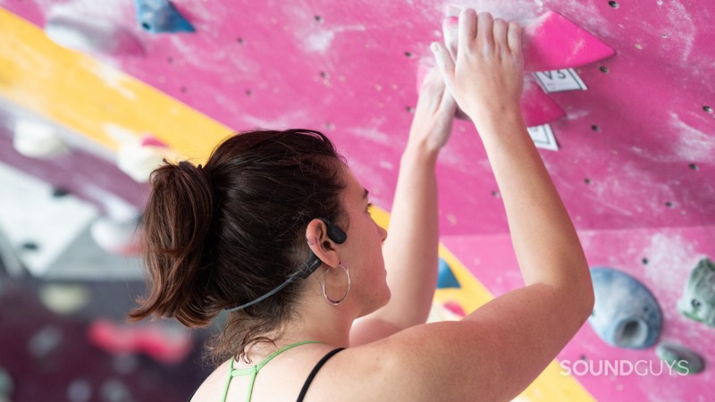 A woman wears the Aftershokz Aeropex bone conduction headphones as she begins to rock climb in a gym.